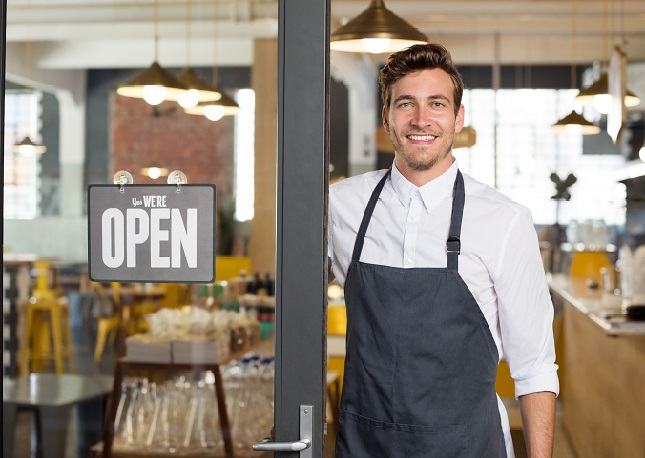 Young bakery owner in doorway with OPEN sign.