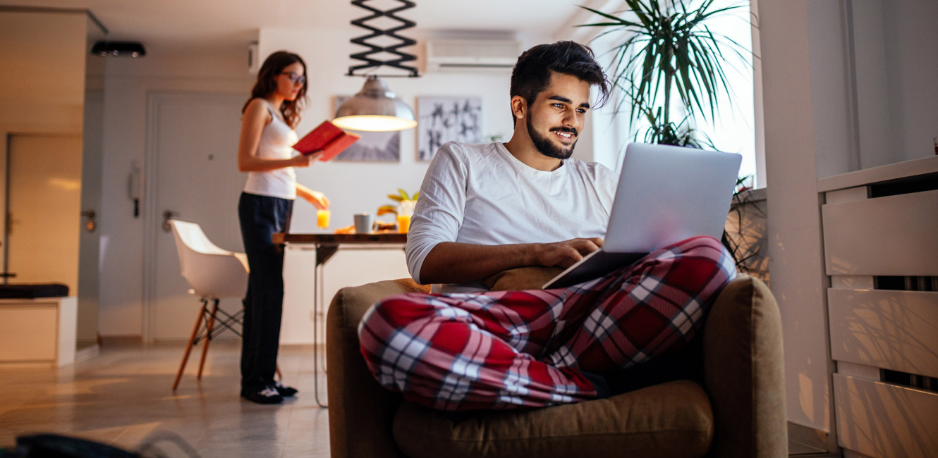 Man in pajamas at home sitting with laptop computer while woman reads a book standing up in background.