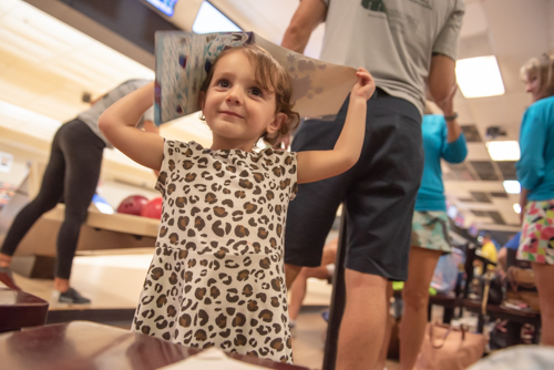 Little girl puts book on her head and smiles while other people talk in the background